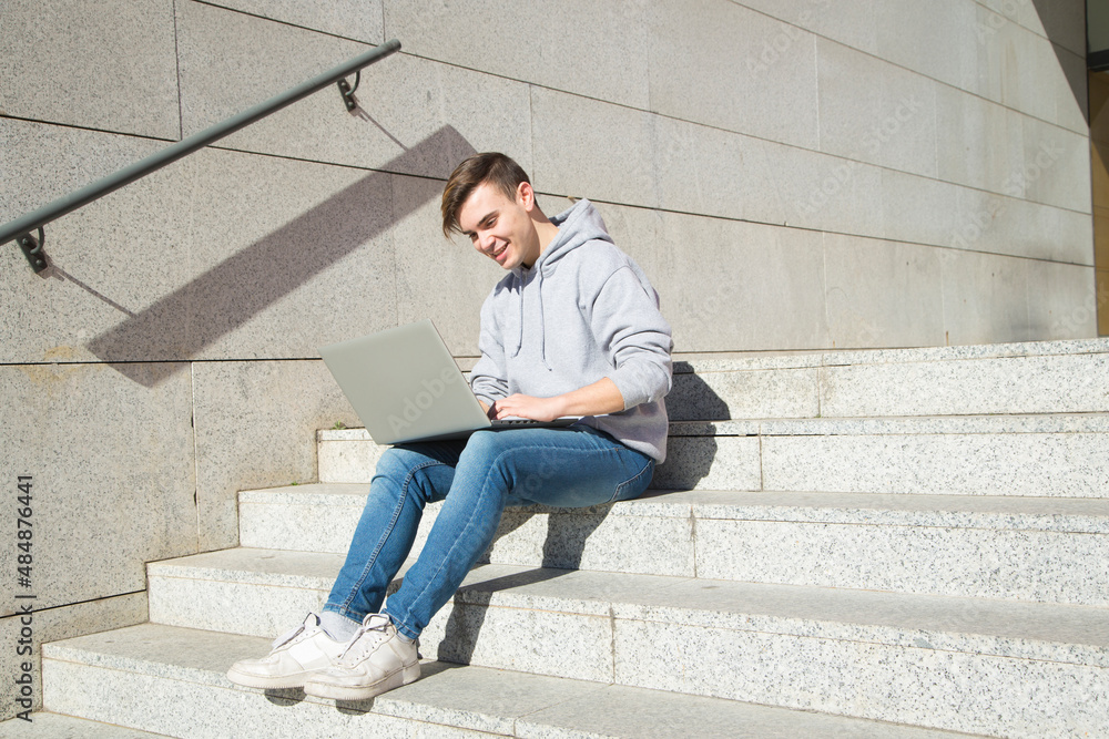 young male college student smiling while sitting on a staircase communicating with a laptop computer young male college student smiling while sitting on a staircase communicating with a laptop computer