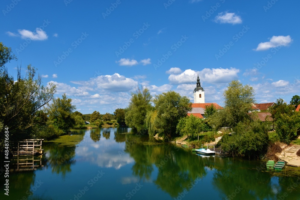 View of Krka river at Kostanjevica na Krki in Dolenjska, Slovenia with a reflection of the sky and trees in water and a church tower rising above the foliage