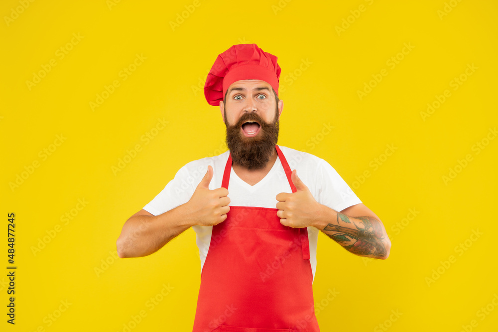Surprised man in toque and apron giving double thumbs yellow background, cook