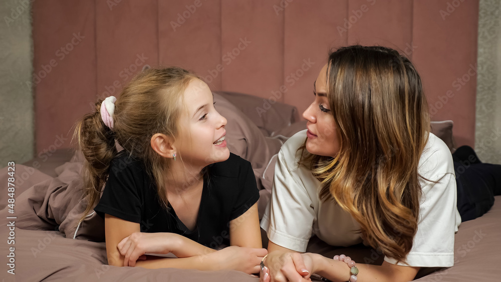 Φωτογραφία Mother and teen daughter in black and white t-shirts hug and kiss lying on bellies on soft bed covered with warm blanket smiling at home, close view