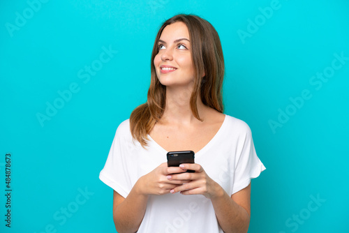 Young caucasian woman isolated on blue background using mobile phone and looking up