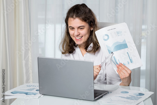Disabled woman freelancer with infantile cerebral paralysis talks on videocall via laptop with manager showing project paper against window at home.