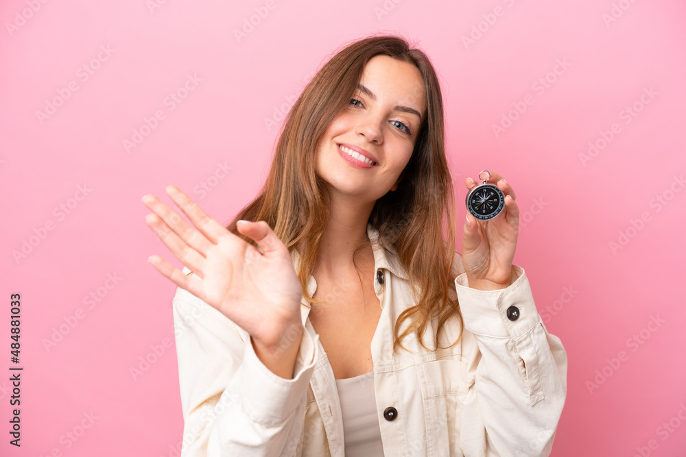 Young caucasian woman holding compass isolated on pink background saluting with hand with happy expression Young caucasian woman holding compass isolated on pink background saluting with hand with happy expression