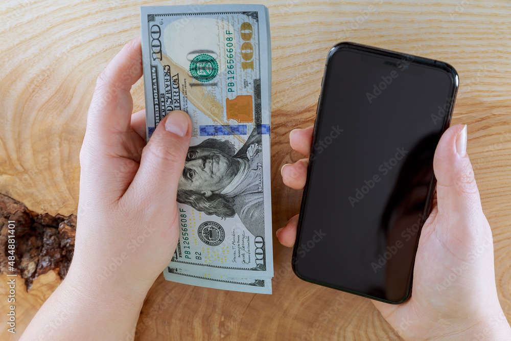 A business woman holds a mobile phone and one hundred dollar bills in her hands on a wooden table background. A business woman holds a mobile phone and one hundred dollar bills in her hands on a wooden table background.