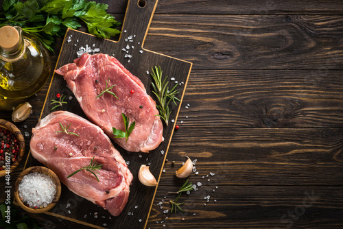 Fresh pork steaks with herbs and spices on dark board at wooden table Top view image with copy space.