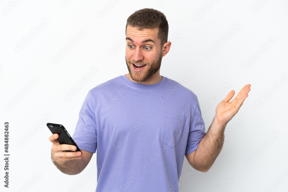 Young handsome caucasian man isolated on white background looking at the camera while using the mobile with surprised expression Young handsome caucasian man isolated on white background looking at the camera while using the mobile with surprised expression