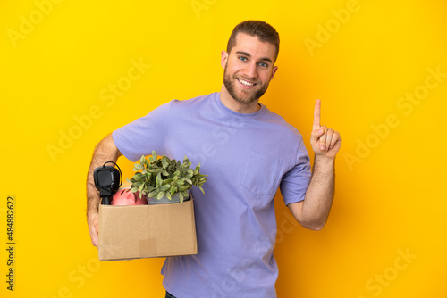 Young caucasian making a move while picking up a box full of things isolated on yellow background showing and lifting a finger in sign of the best