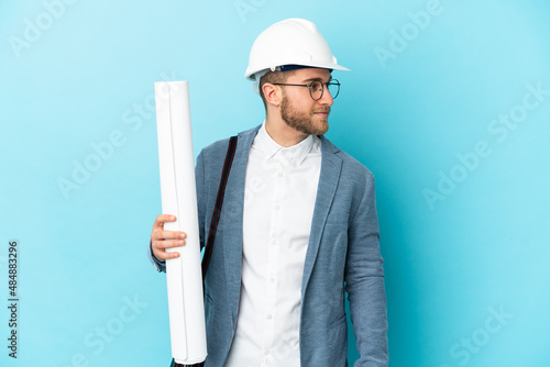 Young architect man with helmet and holding blueprints over isolated background looking to the side photo