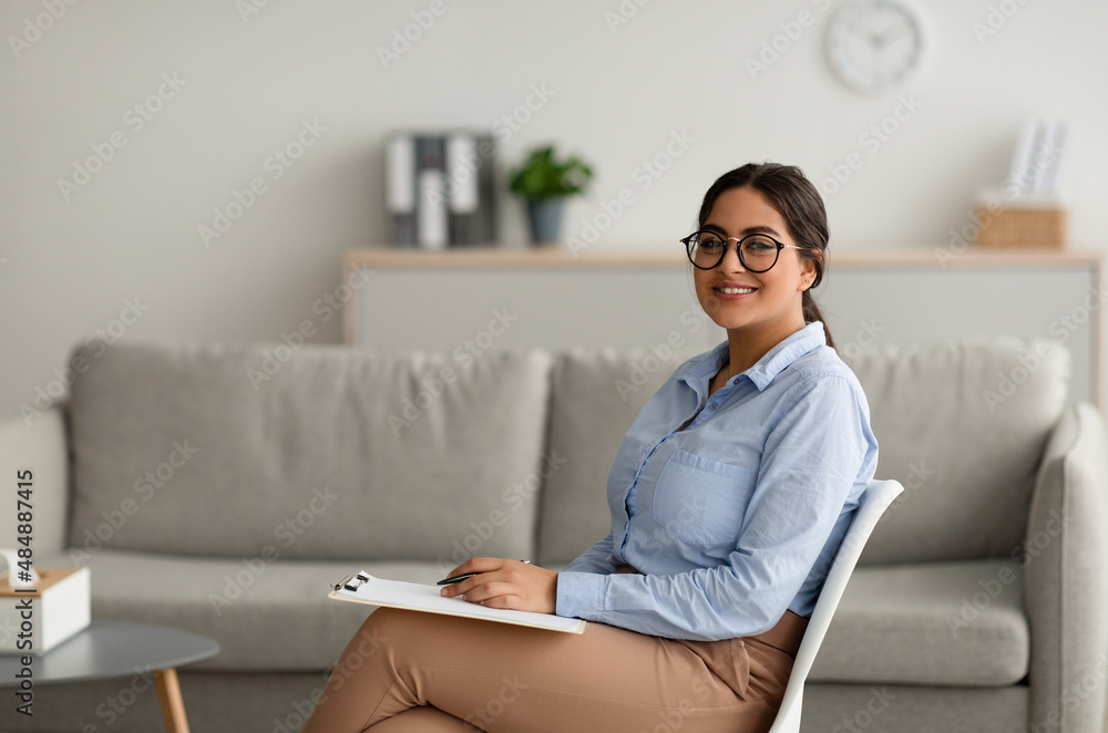 Confident arab female psychologist writing in clipboard, sitting at modern office and smiling at camera, free space - obrazy, fototapety, plakaty 