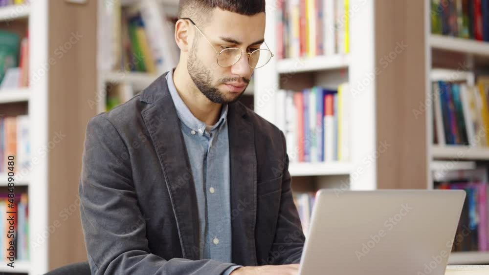 Bearded man with eyeglasses wearing jacket browsing internet on laptop in library, doing research and making notes on paper, camera tilt down, bookcases on blurred background. Concept of education Bearded man with eyeglasses wearing jacket browsing internet on laptop in library, doing research and making notes on paper, camera tilt down, bookcases on blurred background. Concept of education