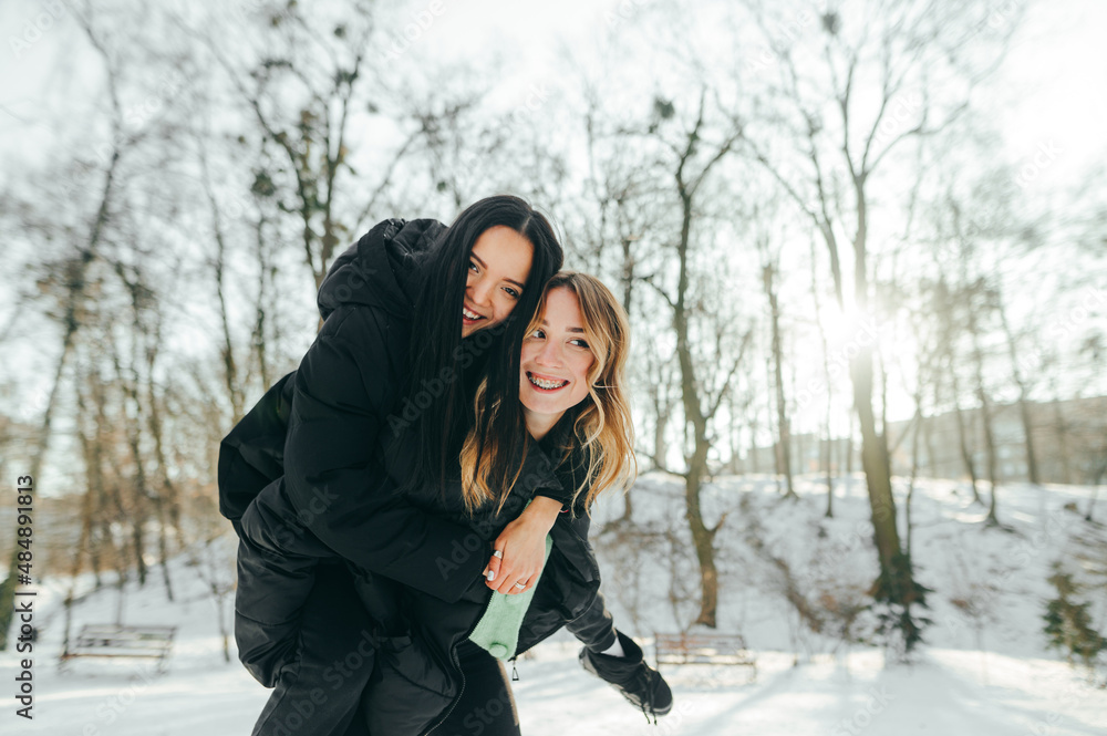 Two cheerful girls girlfriends having fun on a walk in winter park, woman wearing girlfriend on her back with a smile on her face. Two cheerful girls girlfriends having fun on a walk in winter park, woman wearing girlfriend on her back with a smile on her face.
