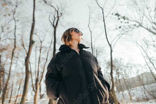 Stylish lady in warm casual clothes stands in a snowy forest at sunset and looks away