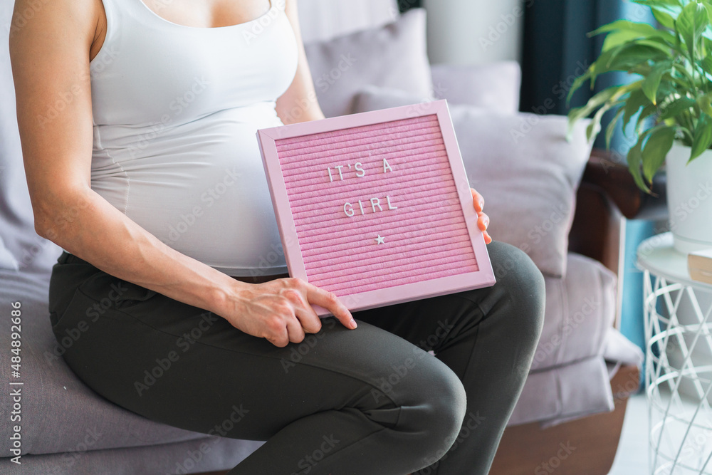 Obraz premium Young unrecognizable pregnant woman in white top and grey trousers sit on grey sofa at home and hold pink board with inscription its a girl. Pregnancy, motherhood, gender role concept.