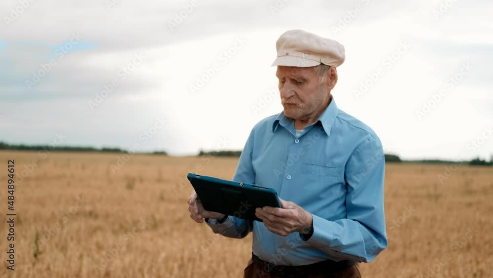 Old gray-haired man in cap standing at field and using tablet device. Senior male farmer tapping and strolling on gadget. Сoncept of modern technology in agriculture. Old gray-haired man in cap standing at field and using tablet device. Senior male farmer tapping and strolling on gadget. Сoncept of modern technology in agriculture.
