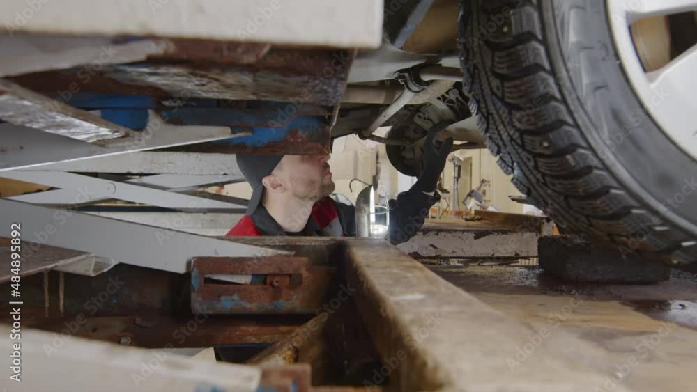 Mechanic standing under lifted car and using lamp while checking bottom in auto service garage Mechanic standing under lifted car and using lamp while checking bottom in auto service garage