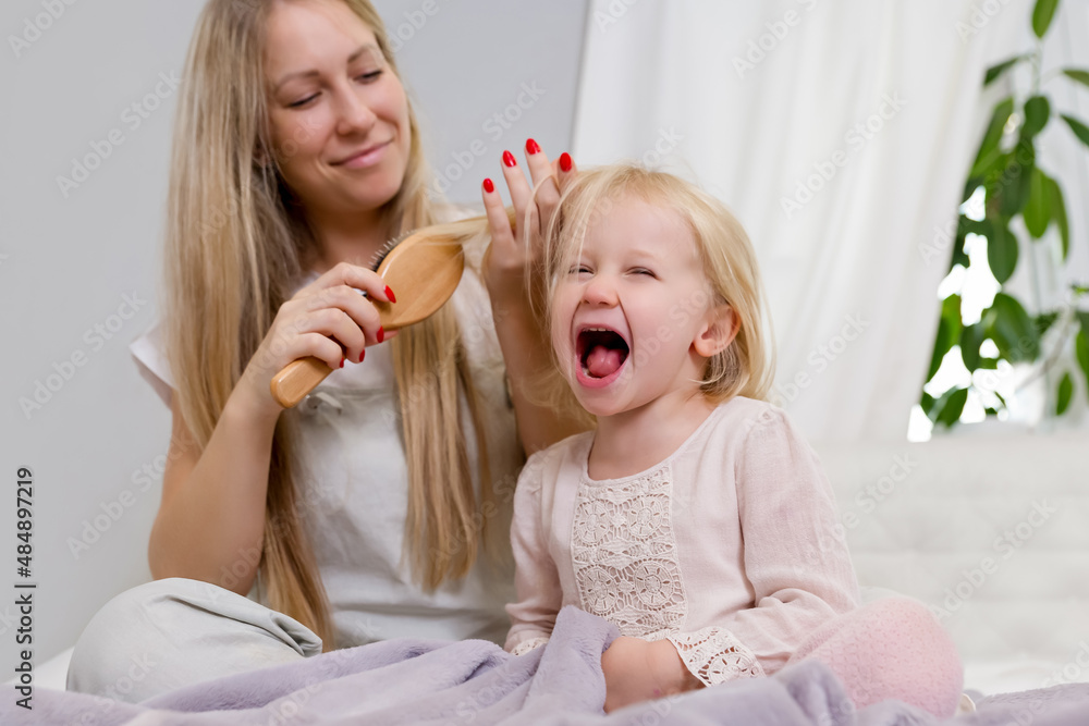 Mother brushes her daughter's blond hair. The little, cheerful girl is laughing and playing with her mother. Family in a light gray interior, minimalist style . Parent playing with child . Naughty kid