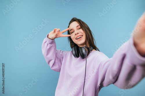 Positive woman in headphones around her neck and in a purple sweatshirt takes a selfie on a blue background with a smile on her face.