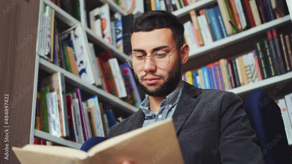 Intelligent man with eyeglasses sitting in book shop and reading book, flipping pages, enjoying interesting plot, bookcases on background, low angle. Concept of education Intelligent man with eyeglasses sitting in book shop and reading book, flipping pages, enjoying interesting plot, bookcases on background, low angle. Concept of education