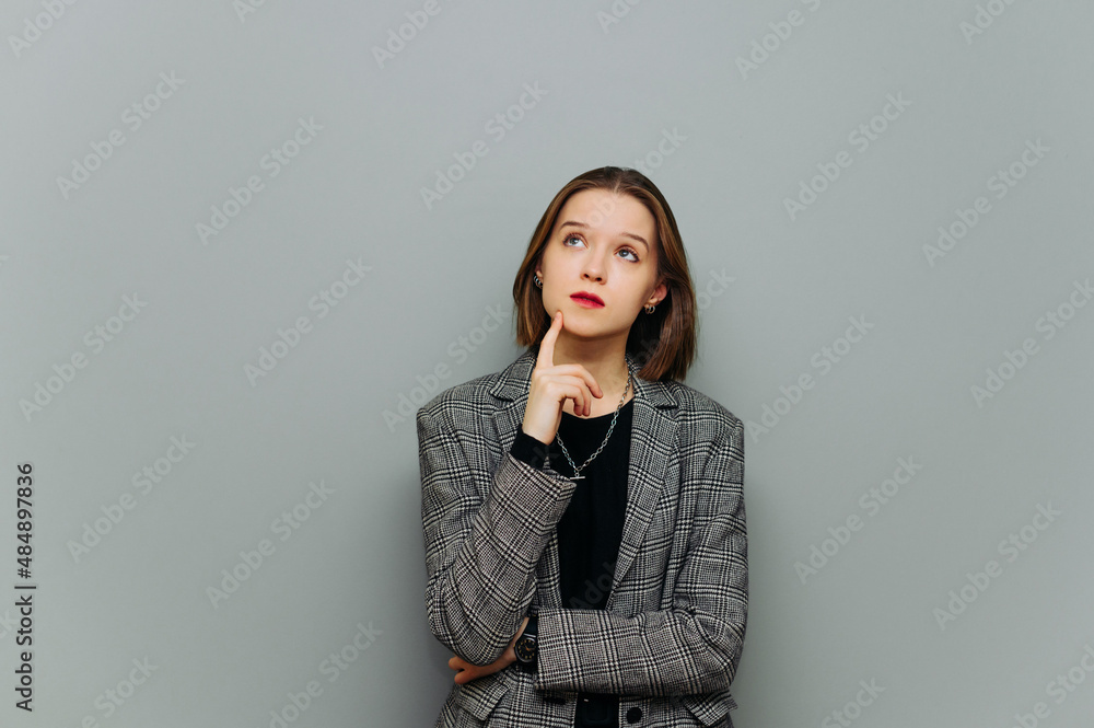Portrait of a pensive young business woman in a jacket, standing on a gray background, looking up and thinking. Portrait of a pensive young business woman in a jacket, standing on a gray background, looking up and thinking.
