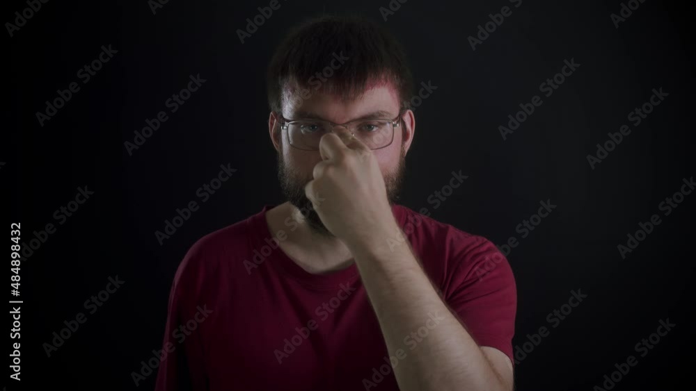 A bearded man wearing glasses and a burgundy T-shirt with a confident look adjusts his glasses and makes a gesture that he is watching you against a black monochromatic background. Front view, slowmo A bearded man wearing glasses and a burgundy T-shirt with a confident look adjusts his glasses and makes a gesture that he is watching you against a black monochromatic background. Front view, slowmo