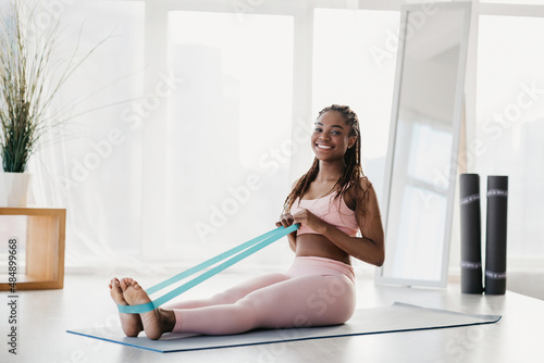 Full length of beautiful African American woman exercising with elastic band on yoga mat during home workout