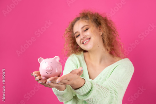 A cute young girl with curly red hair holds a piggy bank, a pink piglet in her hands. The concept of wealth and safety of money.