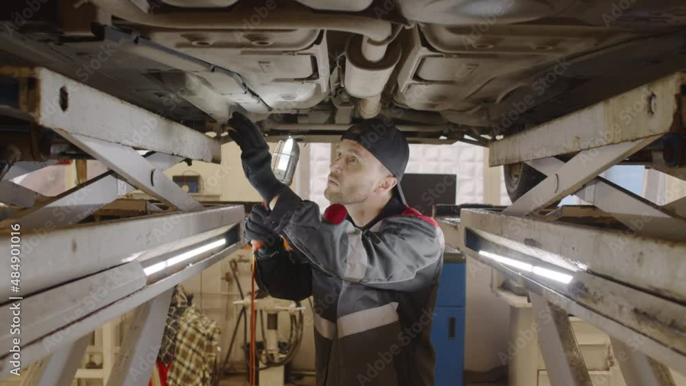 Mechanic walking under lifted car and inspecting car bottom with lamp while working in auto service garage Mechanic walking under lifted car and inspecting car bottom with lamp while working in auto service garage