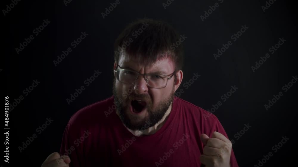 A bearded man wearing glasses and a burgundy T-shirt screams and clenches his fists against a black monochromatic background. Front view, slowmo. A bearded man wearing glasses and a burgundy T-shirt screams and clenches his fists against a black monochromatic background. Front view, slowmo.
