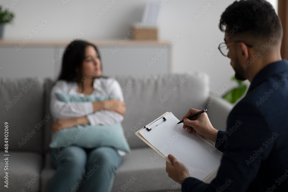 Sad young arab woman hugging pillow and talking to psychologist, focus on psychiatrist taking notes in clipboard Sad young arab woman hugging pillow and talking to psychologist, focus on psychiatrist taking notes in clipboard