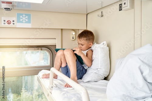 Blond little boy plays online games on tablet sitting and leaning on soft pillow on top shelf in train compartment in morning