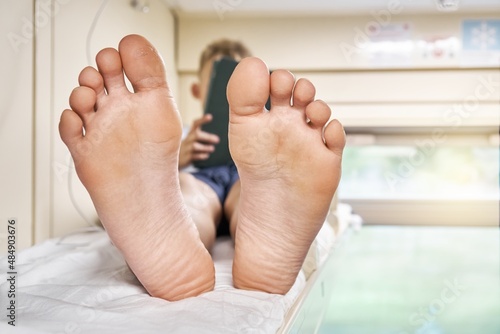 Photography Bare feet of schoolboy lying on top rack bed with white bedding and playing game