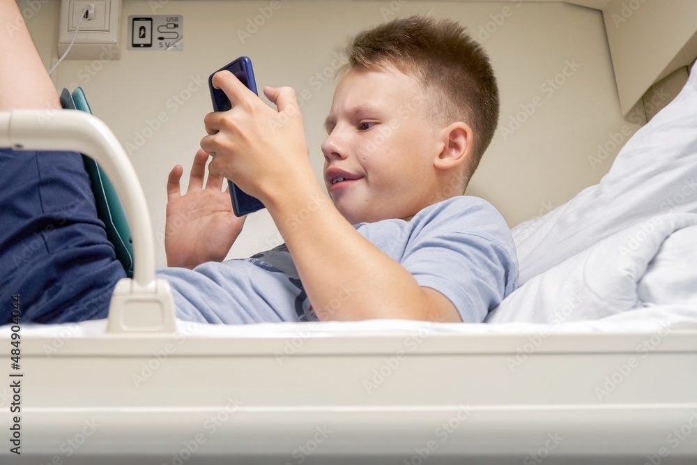 Teen boy with short blond hair reads electronic book with smartphone lying on white bedding on train car upper shelf close low angle shot