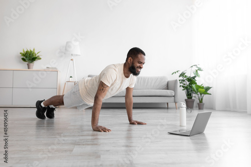 Happy millennial black bearded athlete male in white sports clothes makes plank and watches video on pc