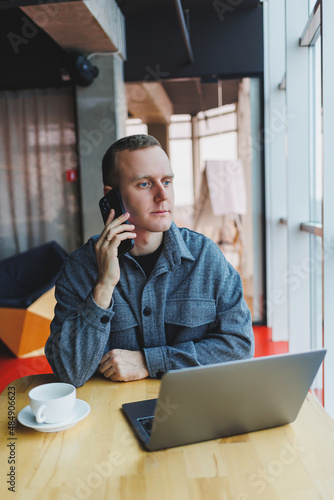 Smiling male entrepreneur in casual wear sitting at table with laptop and talking on smartphone in modern workspace with large window during day