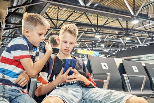 Concentrated blond brothers play games and share interests using smartphones and sitting in waiting hall with armchairs in airport