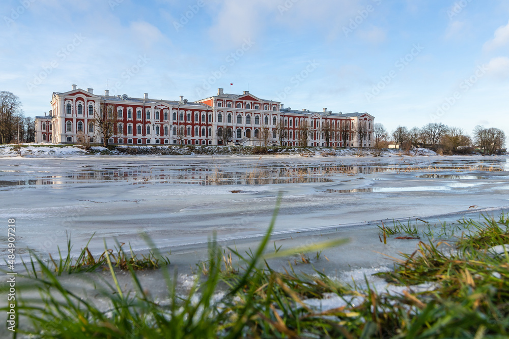 Low angle view to baroque house on the river in winter. Palace in the city of Jelgava, Latvia, Europe. Historical red-white residential building on the frozen river bank.