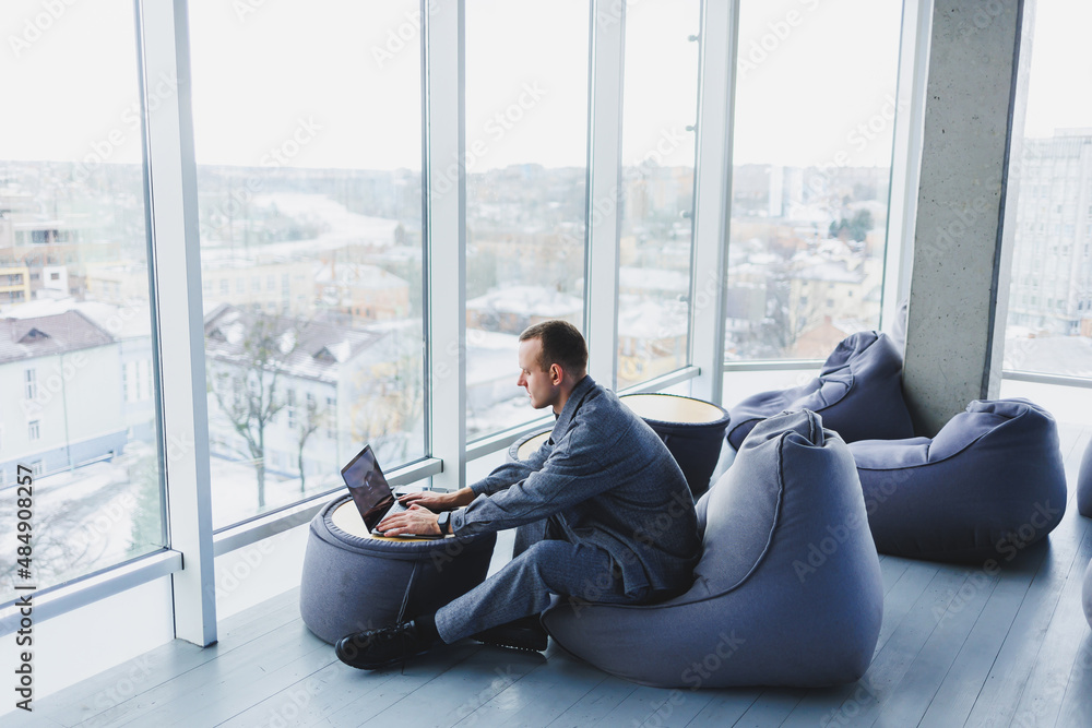 Break. Smiling young man relaxing on bean bag chair while sitting at table and relaxing using laptop, happy businessman leaning back, enjoying work, feeling satisfied