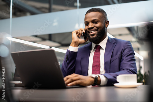 Smiling black manager working on laptop and talking on phone