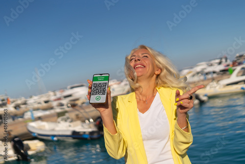 Happy Middle age blonde woman holding a smartphone with a digital health passport in hand against sea marina. New normal travel concept