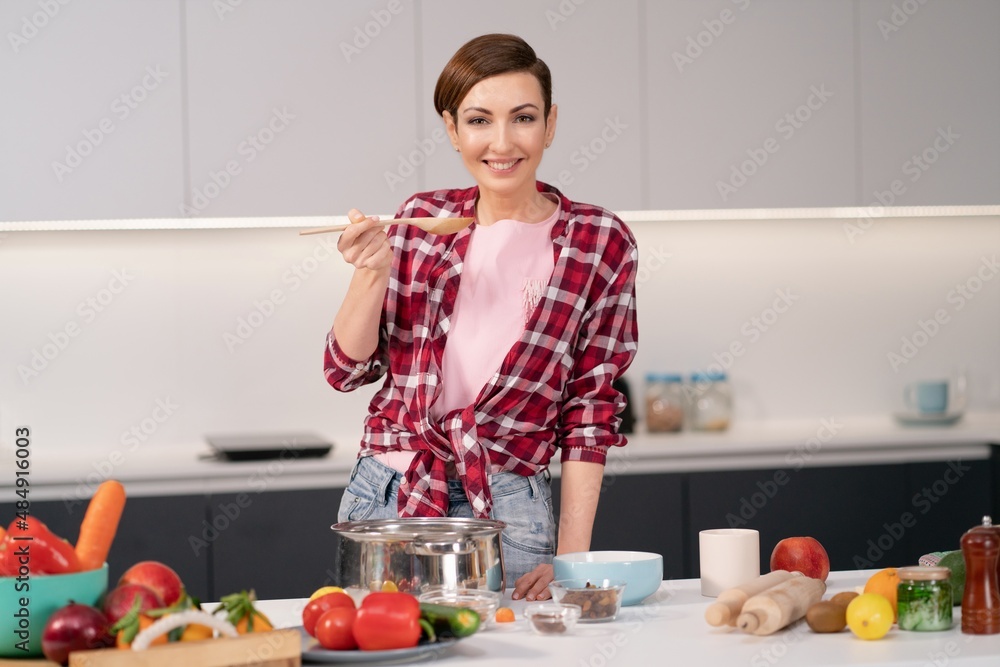 Cheerful housewife in red plaid shirt cooking soup on modern kitchen with lots fresh vegetables on the table in front of her. Smiling woman testing her dish. Chef woman having fun at kitchen Cheerful housewife in red plaid shirt cooking soup on modern kitchen with lots fresh vegetables on the table in front of her. Smiling woman testing her dish. Chef woman having fun at kitchen