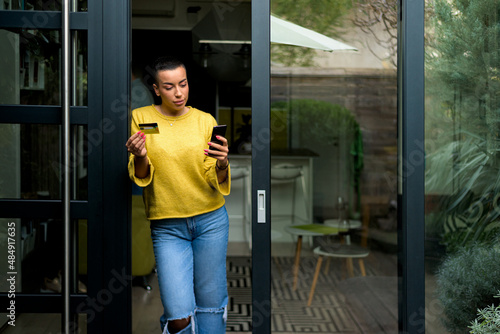 Portrait of casual relaxed woman using phone for online payment on doorstep of her home shopping online from home leaning on door frame using credit card casual entrepreneur doing business from home © Moon Safari