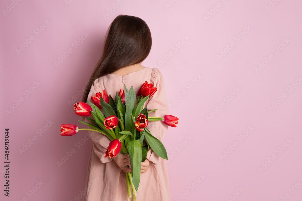 Happy International Women's Day. A cute girl holds a bouquet of red tulips behind her back. Pink background. Space for text.