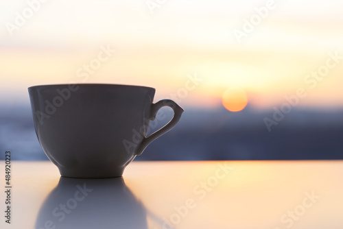 Coffee cup on sunrise background. Fresh start in the morning, view from the window to city in mist