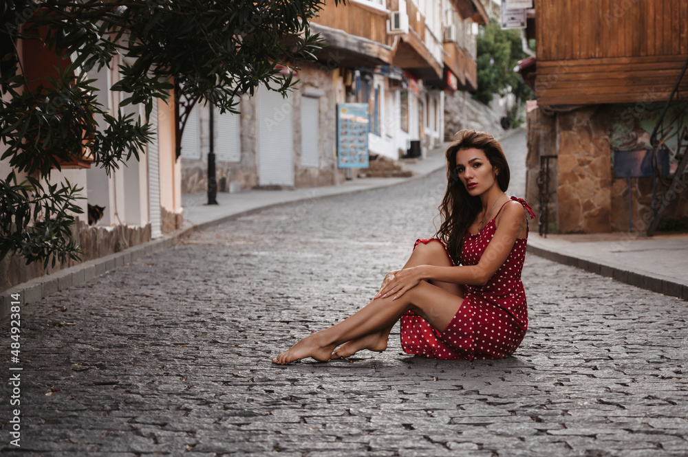 Sexy young Caucasian girl in a red dress posing sitting on a paving stone on a beautiful European street in Italy
