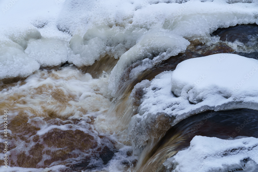 Landscape photo of non-freezing running river with fast current and rapids on its way flowing between ice-covered white banks against backdrop of snowy forest in distance on gloomy cold winter day Landscape photo of non-freezing running river with fast current and rapids on its way flowing between ice-covered white banks against backdrop of snowy forest in distance on gloomy cold winter day