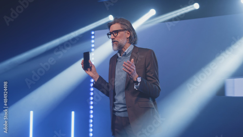 Canvas-taulu A male speaker on stage holding a new smartphone and talking about the character