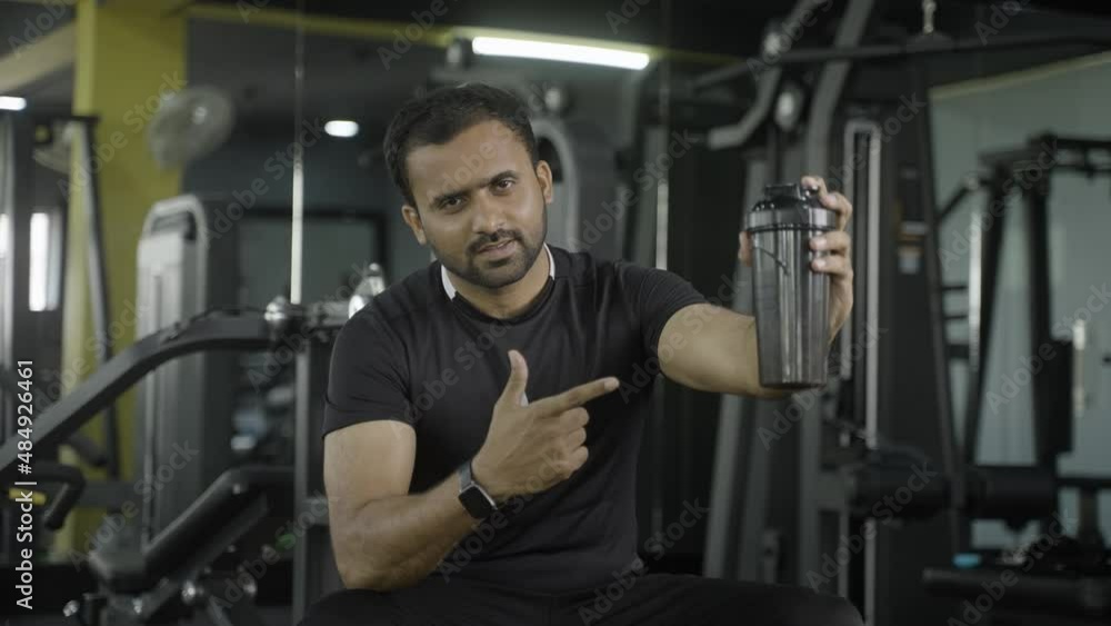 young indian muscle builder with protein powder on shaker bottle showing to camera at gym - concept of advertisment, muscle building and nutrition supplement promotion. young indian muscle builder with protein powder on shaker bottle showing to camera at gym - concept of advertisment, muscle building and nutrition supplement promotion.