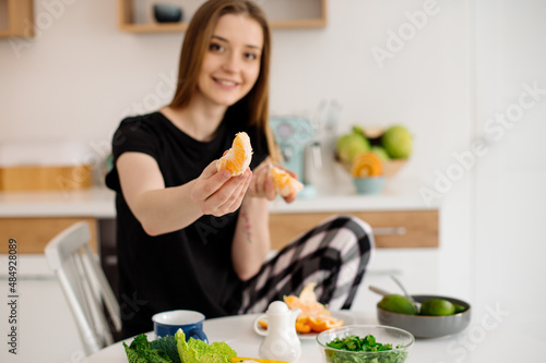 Young beautiful vegetarian girl dressed in pajamas eating fruits and vegetables for breakfast at home in the kitchen