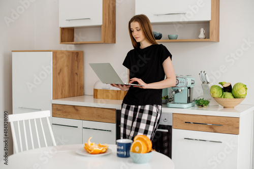 Beautiful young woman in pajamas working with laptop in the kitchen in the morning