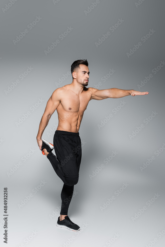 full length of young shirtless sportsman stretching leg and standing with outstretched hand on grey.